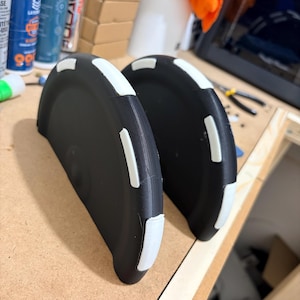 Può includere: Due oggetti neri, semicircolari, con dettagli bianchi. Gli oggetti hanno una forma arrotondata e sono posti uno accanto all'altro. I dettagli bianchi sono rettangolari e posti lungo i bordi degli oggetti neri. Gli oggetti sembrano essere di plastica.