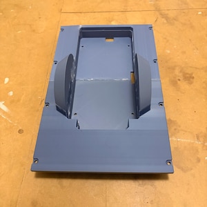 Könnte beinhalten: Ein hellblaues rechteckiges Kunststoffteil mit abgerundeten Kanten und einem zentralen, vertieften Bereich. Zwei halbkreisförmige Strukturen sind an den Seiten angebracht. Das Teil hat mehrere kleine Löcher.