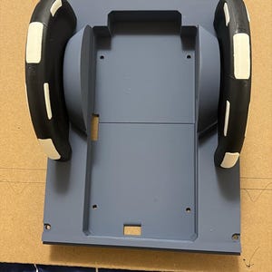 Puede incluir: Una carcasa rectangular de plástico azul mate con un área central empotrada y dos accesorios laterales negros curvados. Cada accesorio negro está acentuado con tres inserciones rectangulares blancas distintas a lo largo de su borde exterior.
