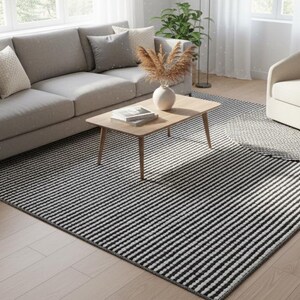 Puede incluir: Un salón moderno con un sofá gris, un sillón beige y una mesa de centro de madera. Una alfombra a rayas blancas y negras cubre el suelo, con una alfombra circular más pequeña a juego. Hay cojines, un jarrón y plantas.