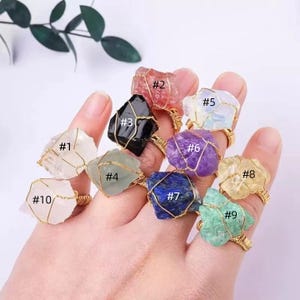May include: A hand displays ten gemstone rings with gold wire settings. The raw, uncut stones are various colors, including clear, red, black, green, purple, blue, and yellow. Each ring is numbered from #1 to #10.