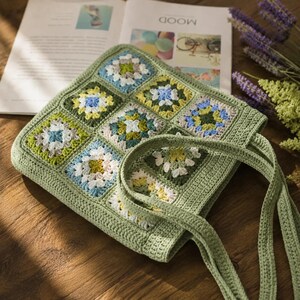 Può includere: Una borsa tote quadrata all'uncinetto composta da diversi quadrati granny nei toni del verde salvia, azzurro, bianco e giallo. La borsa presenta un bordo in tinta unita verde salvia e due manici coordinati all'uncinetto con filato testurizzato.