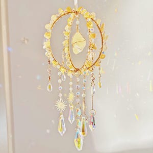 May include: A decorative suncatcher with a celestial theme. It features a gold-colored wire frame adorned with yellow crystals and a large, pale yellow stone in the center. Dangling from the frame are clear crystal pendants and star-shaped charms.
