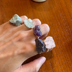 May include: Five rings featuring raw, uncut gemstones wrapped in thin gold-toned wire. The stones include a blue-green crystal, a translucent white crystal, a deep purple amethyst, a dark blue lapis lazuli, and a pale pink rose quartz crystal.