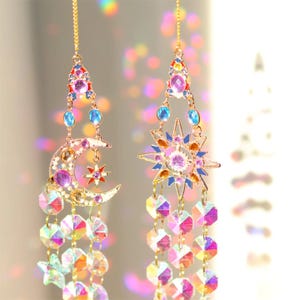 May include: Two celestial-themed hanging ornaments. One features a crescent moon and star, the other a sunburst design. Both have colorful jewel accents and dangling iridescent crystal beads. The ornaments are suspended by delicate gold chains.