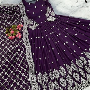 May include: A dark purple dress with detailed silver embroidery and embellishments. The dress has a long, flowing skirt, long sleeves, and a matching scarf. The text "ZARIYAA APPAREL" is visible.