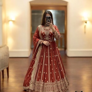 May include: A long, flowing, rust-colored dress with intricate gold embroidery. The dress features a high neckline, long sleeves, and a full skirt. The gold embroidery is concentrated around the neckline, sleeves, and hem. The dress is suitable for formal occasions.