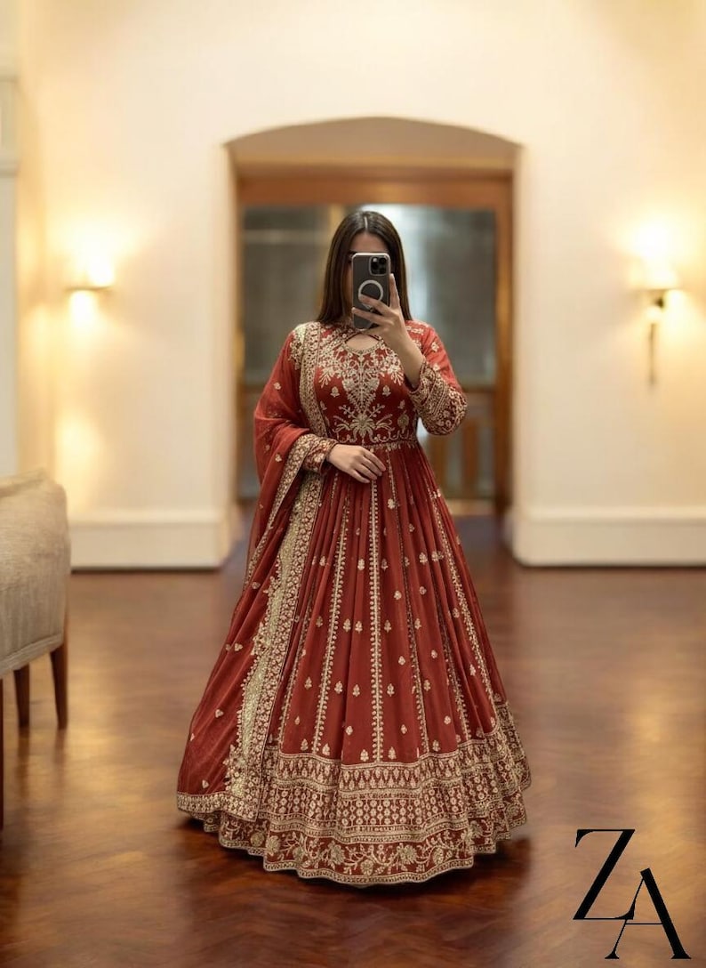May include: A long, flowing, rust-colored dress with intricate gold embroidery. The dress features a high neckline, long sleeves, and a full skirt. The gold embroidery is concentrated around the neckline, sleeves, and hem. The dress is suitable for formal occasions.