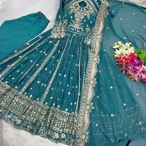 May include: Teal-colored formal dress with intricate silver embroidery and embellishments. The long-sleeved dress features a keyhole neckline and a flowing skirt. A matching scarf and pants are included. The dress is displayed on a wooden hanger.