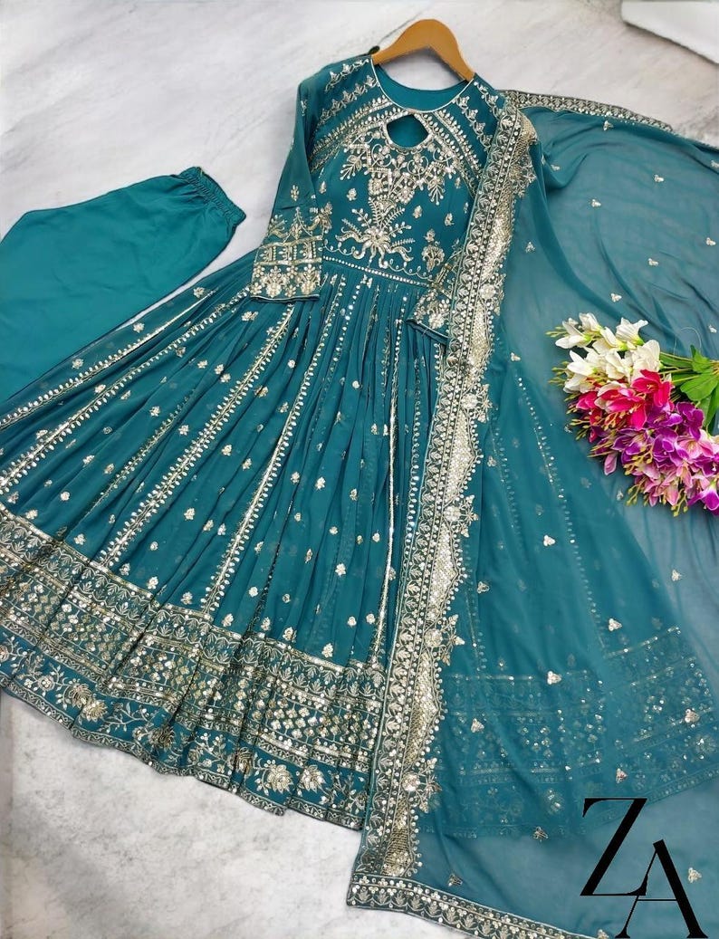 May include: Teal-colored formal dress with intricate silver embroidery and embellishments. The long-sleeved dress features a keyhole neckline and a flowing skirt. A matching scarf and pants are included. The dress is displayed on a wooden hanger.