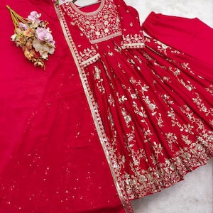 May include: A vibrant red formal dress with intricate gold embroidery and embellishments. The dress features a round neckline, long sleeves, and a flowing skirt. A matching red scarf and pants are included. The dress is displayed on a light background.