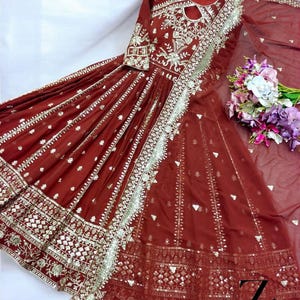 May include: A maroon-colored, long-sleeved dress with intricate silver embroidery and embellishments. The dress features a keyhole neckline and a flowing skirt. A matching sheer scarf and a bouquet of flowers are also present.