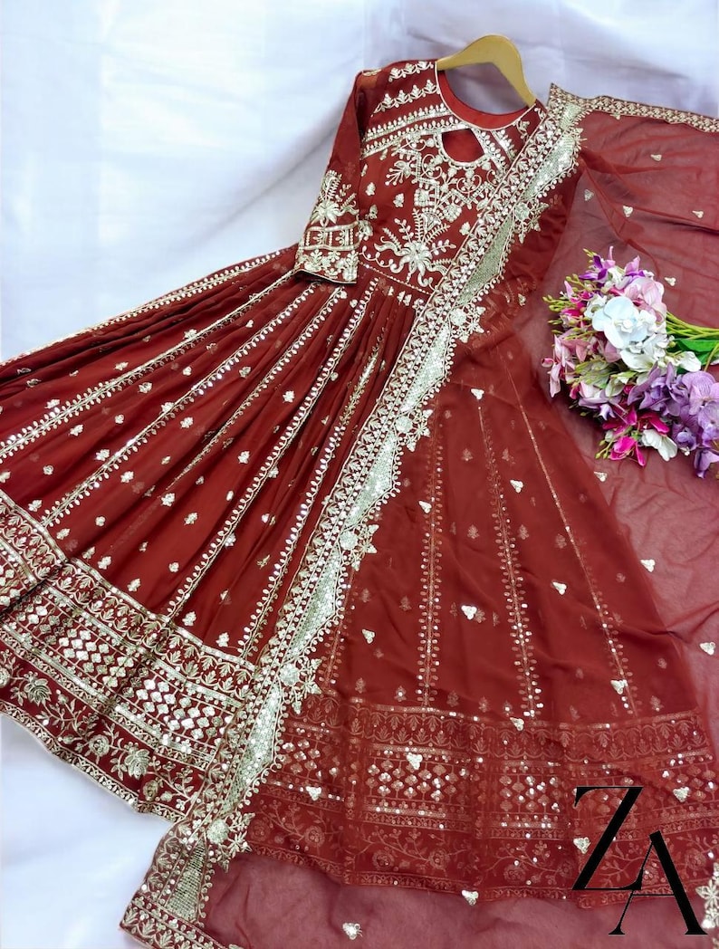 May include: A maroon-colored, long-sleeved dress with intricate silver embroidery and embellishments. The dress features a keyhole neckline and a flowing skirt. A matching sheer scarf and a bouquet of flowers are also present.