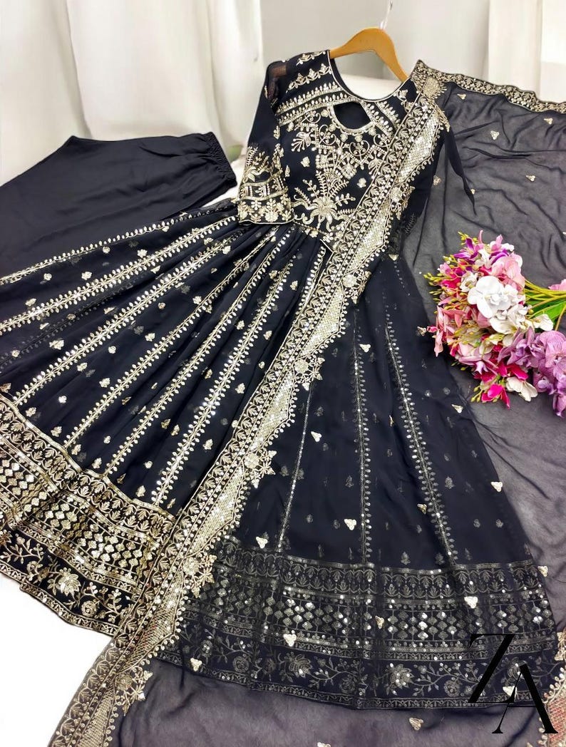 May include: A black formal dress with intricate gold embroidery and embellishments. The dress features a keyhole neckline, long sleeves, and a flowing skirt. A matching black scarf and pants are included. A bouquet of flowers is visible.