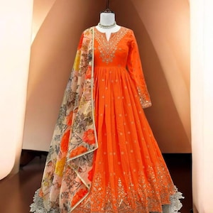 May include: An orange long-sleeved dress with intricate embroidery around the neckline, sleeves, and hem. A floral patterned scarf is draped over the dress. The dress is displayed on a mannequin.