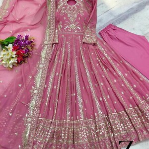May include: A pink, long-sleeved dress with intricate gold embroidery and embellishments. The dress features a flowing skirt and is paired with matching pants and a sheer scarf. A bouquet of flowers is visible on the left.