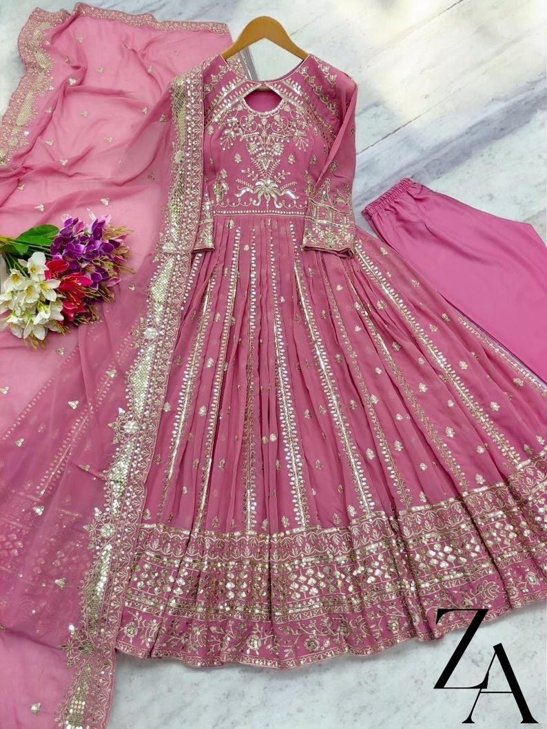 May include: A pink, long-sleeved dress with intricate gold embroidery and embellishments. The dress features a flowing skirt and is paired with matching pants and a sheer scarf. A bouquet of flowers is visible on the left.