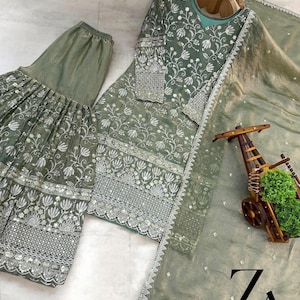 Pu&ograve; includere: Completo ricamato in tre pezzi verde salvia. Il set comprende un top a maniche lunghe, pantaloni a gamba larga e una sciarpa trasparente. Il ricamo floreale &egrave; bianco e argento. L'outfit &egrave; esposto su una gruccia di legno.