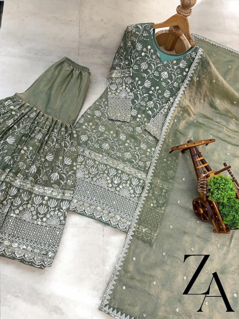 Pu&ograve; includere: Completo ricamato in tre pezzi verde salvia. Il set comprende un top a maniche lunghe, pantaloni a gamba larga e una sciarpa trasparente. Il ricamo floreale &egrave; bianco e argento. L'outfit &egrave; esposto su una gruccia di legno.