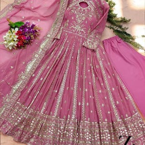 May include: A pink, long-sleeved dress with intricate gold embroidery and embellishments. The dress features a keyhole neckline and a flowing skirt. A matching pink scarf and pants are included. A bouquet of flowers is placed near the dress.