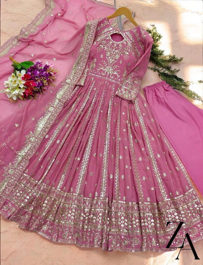 May include: A pink, long-sleeved dress with intricate gold embroidery and embellishments. The dress features a keyhole neckline and a flowing skirt. A matching pink scarf and pants are included. A bouquet of flowers is placed near the dress.