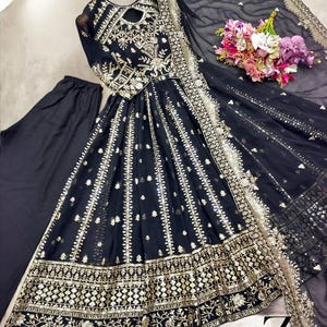 May include: A black embroidered formal dress with gold accents, long sleeves, and a keyhole neckline. The dress includes matching black pants and a sheer black scarf with gold trim. A bouquet of flowers rests on the side.