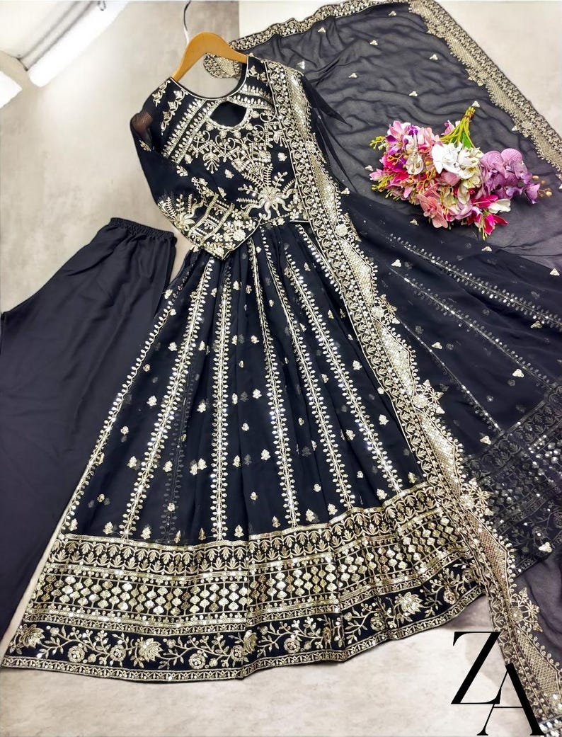 May include: A black embroidered formal dress with gold accents, long sleeves, and a keyhole neckline. The dress includes matching black pants and a sheer black scarf with gold trim. A bouquet of flowers rests on the side.