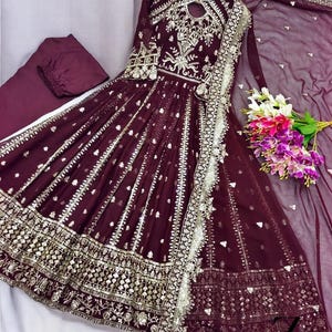 May include: A burgundy formal dress with intricate silver embroidery and embellishments. The dress features a flowing skirt, long sleeves, and a matching dupatta. A bouquet of flowers rests near the dress.