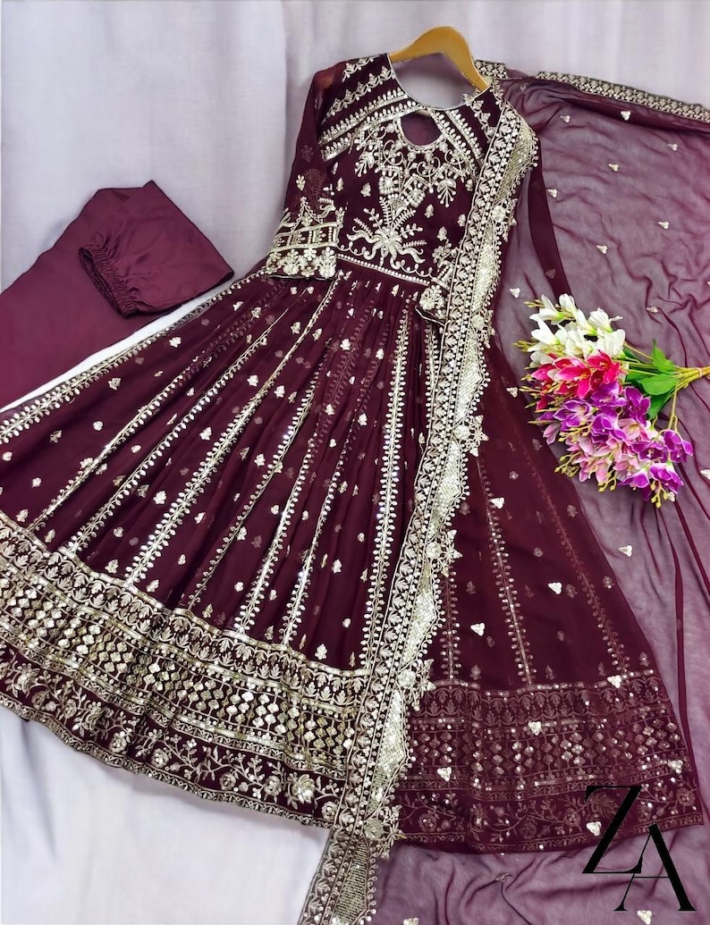 May include: A burgundy formal dress with intricate silver embroidery and embellishments. The dress features a flowing skirt, long sleeves, and a matching dupatta. A bouquet of flowers rests near the dress.