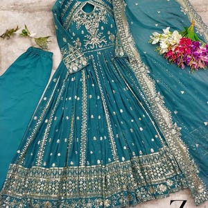 May include: Teal-colored formal dress with intricate silver embroidery and embellishments. The dress features long sleeves, a keyhole neckline, and a flowing skirt. A matching scarf and pants are included. The dress is displayed with a bouquet of flowers.