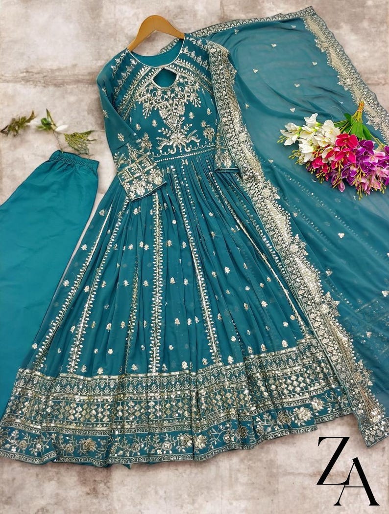 May include: Teal-colored formal dress with intricate silver embroidery and embellishments. The dress features long sleeves, a keyhole neckline, and a flowing skirt. A matching scarf and pants are included. The dress is displayed with a bouquet of flowers.