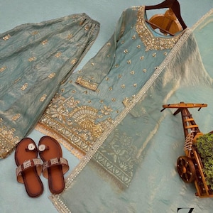 Puede incluir: Un conjunto salwar kameez bordado en azul claro con pantalones a juego y una dupatta transparente. El conjunto presenta adornos dorados y se combina con sandalias marrones. El conjunto se muestra sobre una superficie azul claro.