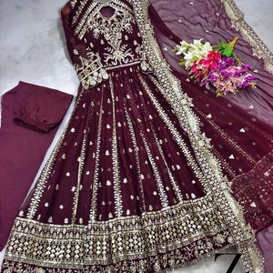 May include: A burgundy formal dress with intricate gold embroidery and embellishments. The dress features a keyhole neckline, long sleeves, and a flowing skirt. A matching scarf and pants are included. A bouquet of flowers rests on the side.
