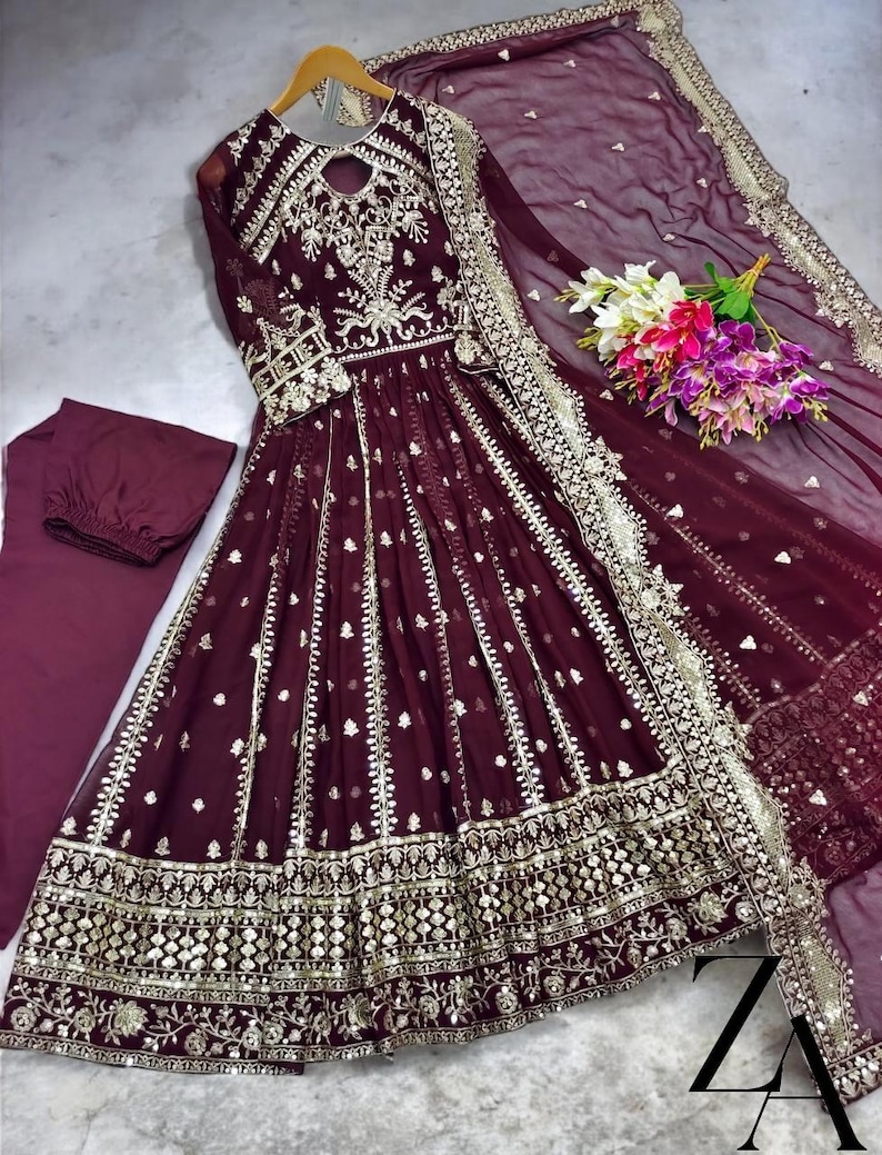 May include: A burgundy formal dress with intricate gold embroidery and embellishments. The dress features a keyhole neckline, long sleeves, and a flowing skirt. A matching scarf and pants are included. A bouquet of flowers rests on the side.