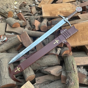 May include: A steel longsword with a brown leather-wrapped grip and a circular pommel featuring a red cross emblem. The blade has a central fuller and an inscription. It is paired with a brown leather scabbard adorned with two gold-coloured crosses.