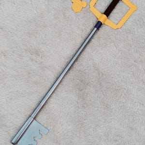 May include: A Kingdom Hearts Keyblade prop featuring a long silver metal shaft, a yellow handguard with a black grip, and a silver key-shaped tip. A silver chain is attached to the handguard, ending in a yellow charm shaped like a mouse head.