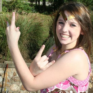 May include: A young person with brown hair is wearing a pink and brown patterned tank top and a gold headband with a red jewel. They are making a rock and roll hand gesture with both hands.