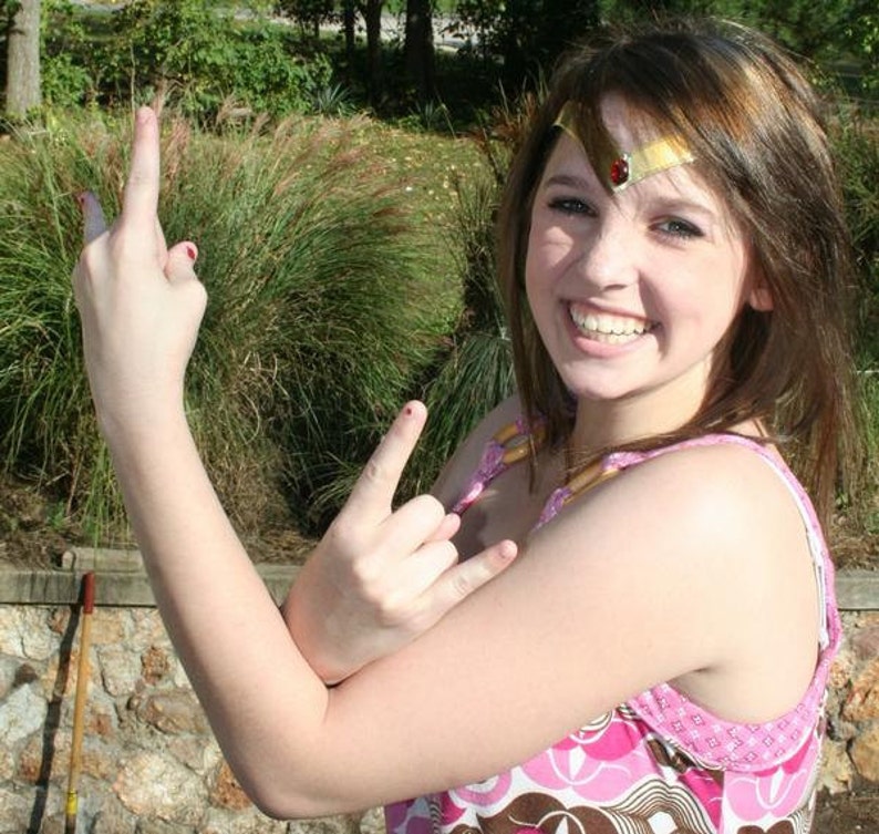 May include: A young person with brown hair is wearing a pink and brown patterned tank top and a gold headband with a red jewel. They are making a rock and roll hand gesture with both hands.