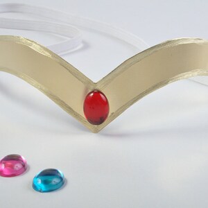 May include: A gold-colored tiara with a red gem in the center and two extra gems, one pink and one blue, on a white surface.