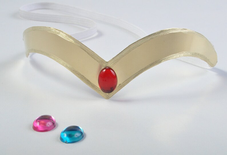 May include: A gold-colored tiara with a red gem in the center and two extra gems, one pink and one blue, on a white surface.