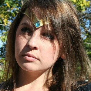 May include: A woman with long brown hair wearing a gold headband with a blue gemstone. The headband is a popular accessory for cosplay and costume parties.