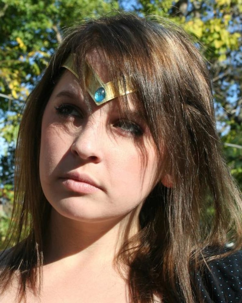 May include: A woman with long brown hair wearing a gold headband with a blue gemstone. The headband is a popular accessory for cosplay and costume parties.