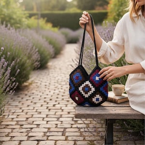Peut inclure: Un sac fourre-tout en crochet fait à la main avec un motif géométrique dans les tons de bleu, rouge, blanc et marron. Le sac a des anses noires et une bordure noire. Le sac est présenté devant un jardin de lavande.