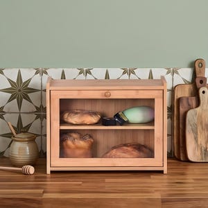 May include: A natural bamboo bread bin with a glass door, displaying two shelves with loaves of bread. A small wooden knob opens the door. The bread bin is on a wooden surface, with cutting boards and a honey pot in the background.