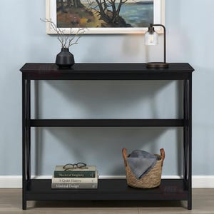 May include: Black console table with three shelves. The top shelf holds a black vase with branches, and a small lamp. The bottom shelf has books and a woven basket. The table has an X-shaped design on the sides.