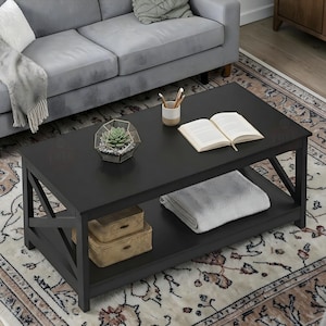 May include: A black rectangular coffee table with an open lower shelf. The table has an X-shaped design on the sides. A succulent terrarium, open book, and decorative boxes are on the table. A grey blanket is on the lower shelf.