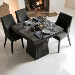 May include: A contemporary dining set featuring a dark grey square table with a pedestal base, set for four. The table is surrounded by four grey upholstered chairs. Plates, cutlery, and glassware are arranged on the table. A fireplace with a burning fire is in the background.