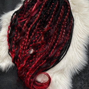 Puede incluir: Un conjunto de rastas negras y rojas con una mezcla de texturas trenzadas y retorcidas. Las rastas están adornadas con cintas rojas, cuentas y un adorno decorativo. El cabello se presenta sobre una superficie de piel sintética blanca.