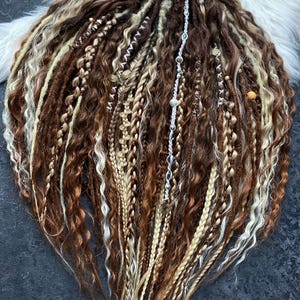 May include: A collection of brown, blonde, and light brown dreadlocks, some with decorative beads and charms. The dreadlocks are arranged in a fan shape, with a variety of textures and styles, including braids and twisted sections. The hair is tied with a brown ribbon.