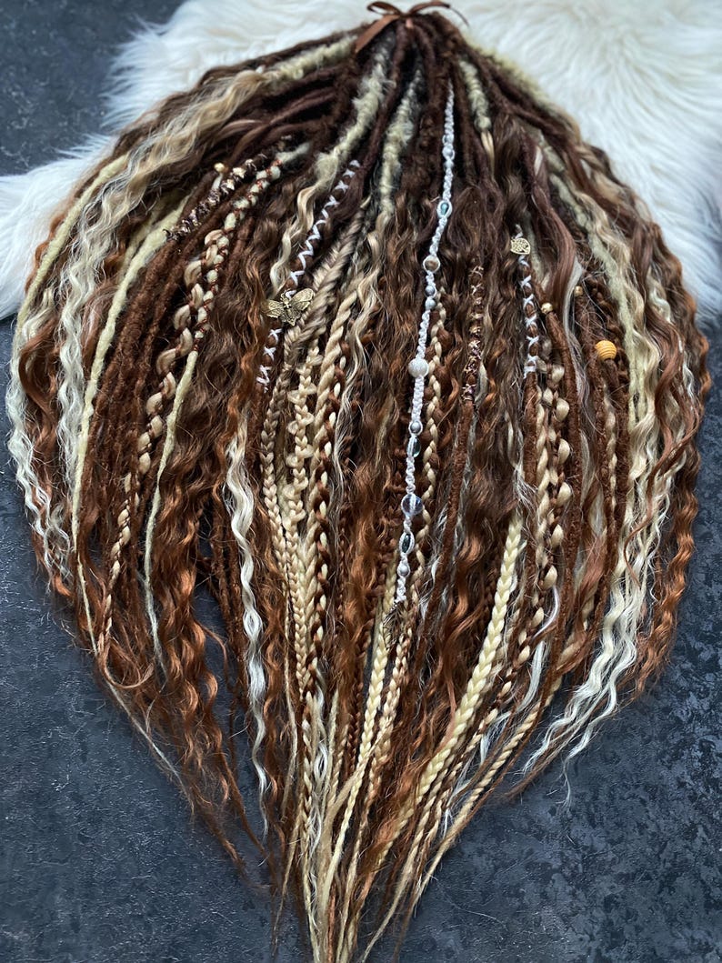 May include: A collection of brown, blonde, and light brown dreadlocks, some with decorative beads and charms. The dreadlocks are arranged in a fan shape, with a variety of textures and styles, including braids and twisted sections. The hair is tied with a brown ribbon.